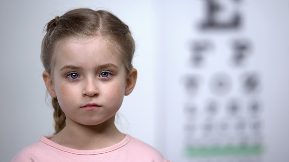 Lille pige med synsproblemer står foran en synstavle og ser ind i kameraet